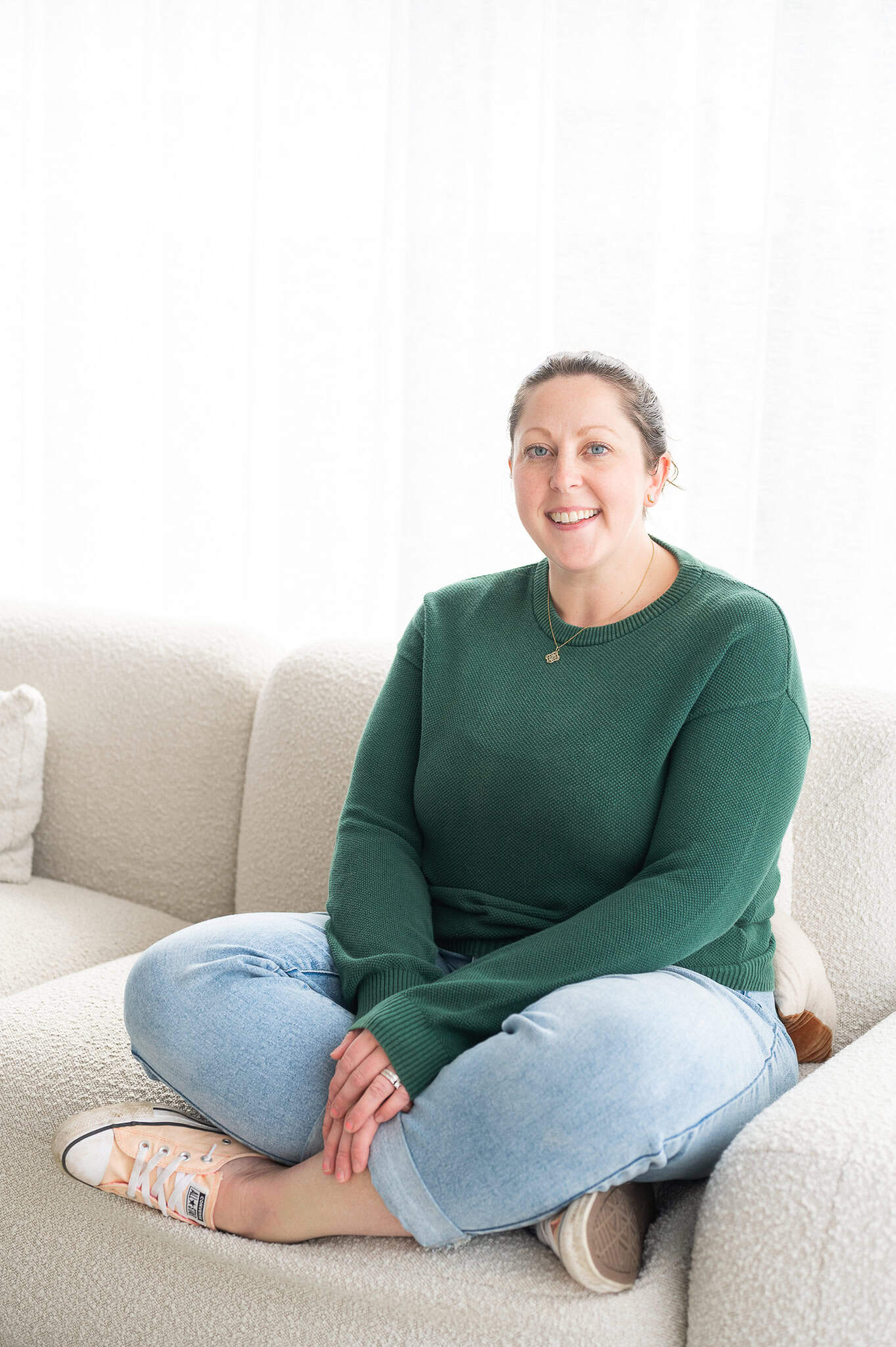 Dacia-10 Relaxed woman sitting on a white sofa, wearing casual green sweater and jeans, smiling at the camera, in a bright and airy living room setting.